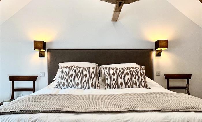 A tasteful neutral coloured bedroom with exposed beams and large bed with decorative cushions and throw, with an antique chair to each side of the bed
